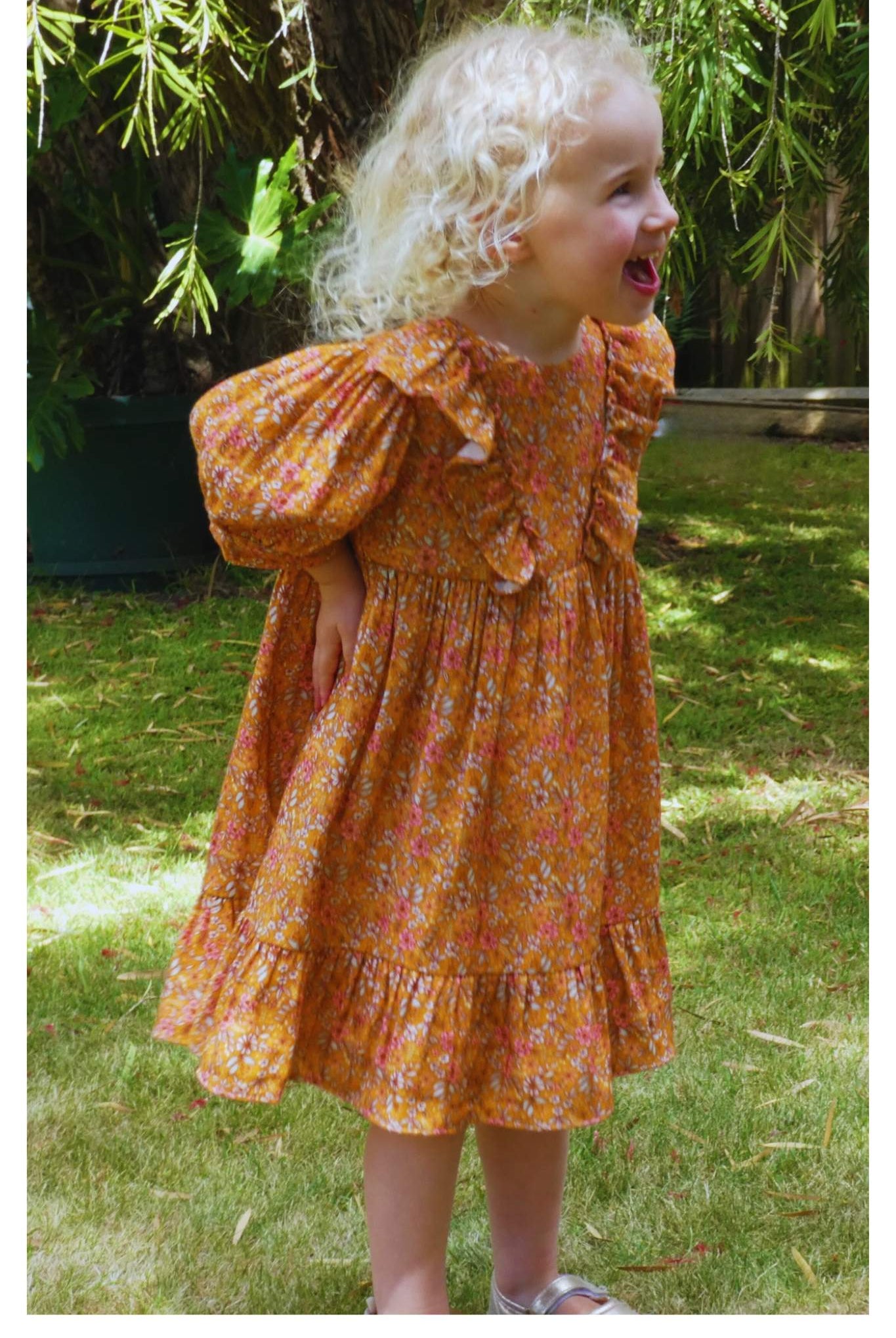 A child wearing a vintage style print dress in orange/mustard/cream with a gathered yoke, diagonal fabric frills on the bodice, a zipper at the back, and frilled hemline. The dress features longer style puff sleeves and is gathered onto a loose cuff.
