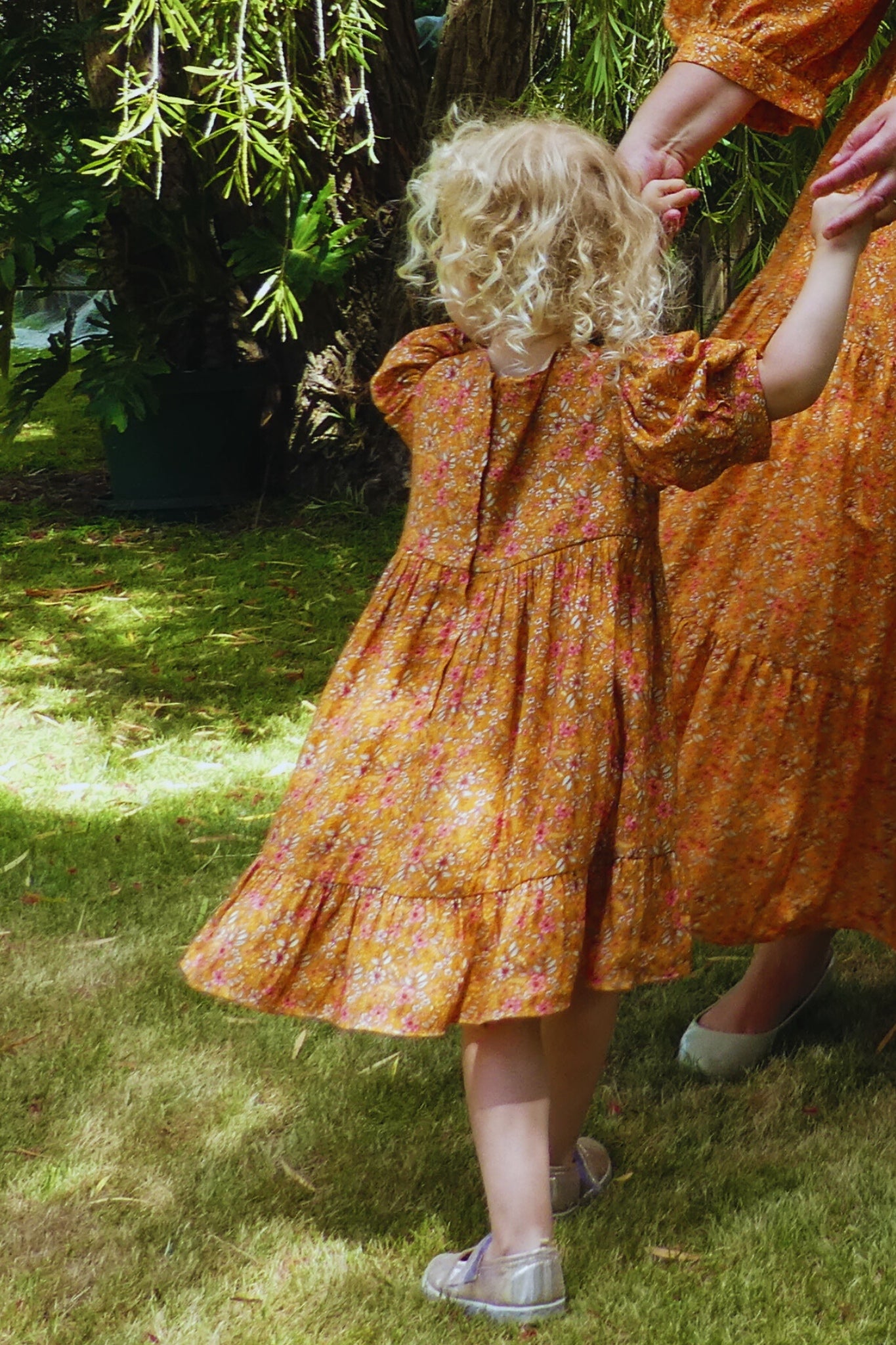 Child and adult wearing matching orange floral dresses outdoors on a grassy area.